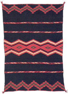 There is a photograph of a woven black blanket with lines and zigzag patterns woven across it using red, black, white, and green colors. The corners are tufted.