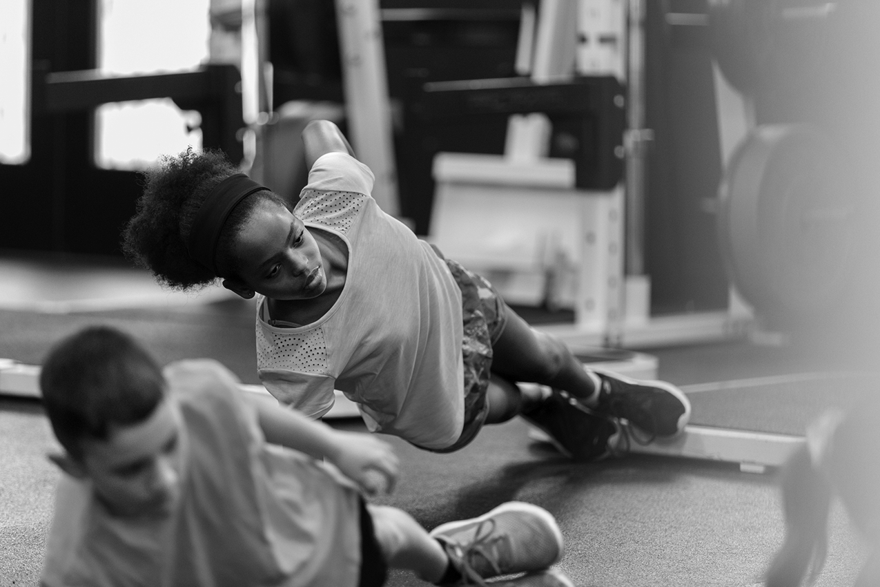 The photo shows two young people with their bodies straight and propped up from a gym floor by their right arms.
